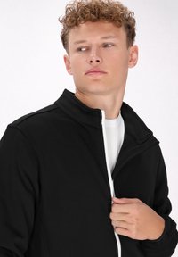 Giacca nera con zip e colletto alto, caratterizzata da dettagli bianchi sulla zip, realizzata in tessuto morbido, mostrata parzialmente aperta.