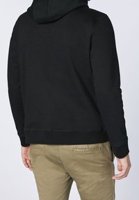 Svart huvtröja med en jämn textur, med ribbad mudd och fåll. Buren med ljusbeiga byxor, som visar baksidan.