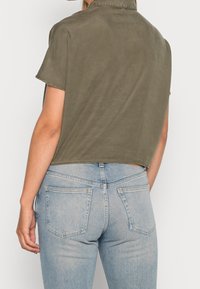 Camicia verde oliva a maniche corte con una vestibilità leggermente ampia, caratterizzata da un orlo dritto e colletto, indossata con jeans azzurri chiari.