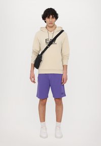 Felpa beige con testo nero, pantaloni corti viola, scarpe da ginnastica bianche e una piccola borsa a tracolla nera. Design semplice e vestibilità rilassata.