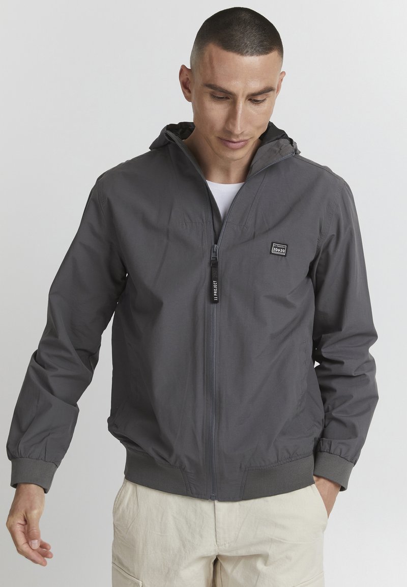 Giacca leggera grigia con cappuccio, polsini elasticizzati e zip. Presenta un piccolo logo sul petto e un design dalla vestibilità comoda.