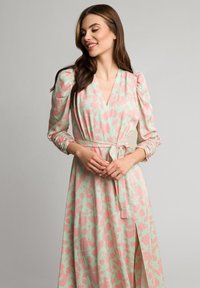 Sukienka w kwiaty w delikatnym zielonym kolorze z różowymi wzorami, z długimi bufkami, dekoltem w serek oraz przewiązanym w pasie z boku rozcięciem.