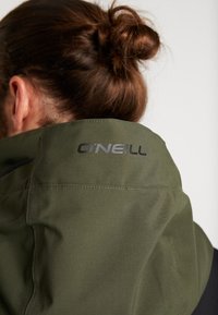 Personne aux cheveux bruns en chignon portant une veste vert olive avec le texte noir "O'NEILL" sur le col arrière.