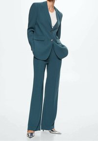 Čistý tyrkysový oblek na míru s jedním knoflíkem na saku, kalhoty s rovnými nohavicemi a hladkou texturou. Doplněno stříbrnými botami se špičatou špičkou.