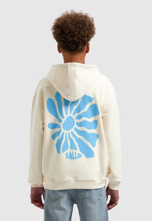 Persoon met krullend haar die een crème hoodie draagt met een grote blauwe abstracte bloem en de tekst "BALLIN" op de achterkant, gecombineerd met lichtblauwe spijkerbroeken.