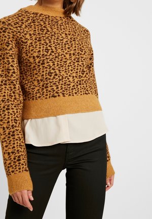 Femme portant un pull en maille marron à motifs avec poignets et ourlet côtelés, par-dessus une blouse blanche et un pantalon noir, cadrée à mi-torso.