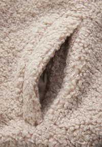 Beige tekstureret stof med en blød, fluffy overflade, der har en fremtrædende lommeåbning. Materialet fremstår hyggeligt og indbydende.