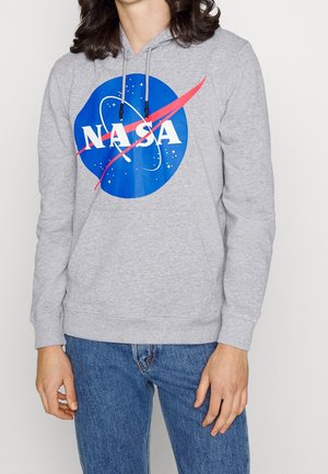 Osoba ubrana w szarą bluzę z kapturem z niebieskim logo NASA i niebieskie dżinsy, stojąca na tle jednolitego białego tła.