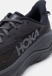 Κοντινό πλάνο μαύρου παπουτσιού τρεξίματος της μάρκας HOKA με υφασμένο επάνω μέρος με υφή, χοντρή επενδυμένη σόλα και γκρι κορδόνια.