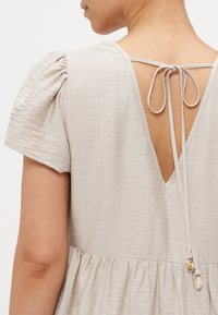 Φωτεινό μπεζ φόρεμα με φουσκωτά μανίκια, v-λαιμόκοψη και κορδόνι. Διαθέτει συγκεντρωμένη μέση, υφασμάτινη υφή και διακοσμητικά στοιχεία σε σχήμα κοχυλιού στο κορδόνι.