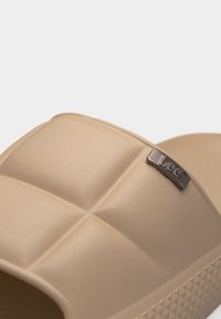 Κοντινό πλάνο μπεζ σανδαλιού με ριγέ λουράκι και μικρό μεταλλικό λογότυπο Lee στο πάνω μέρος.