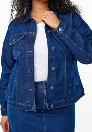 Femme portant une veste et une jupe en denim bleu foncé, tenant le col de la veste, avec une chemise blanche en dessous, sur un fond uni.