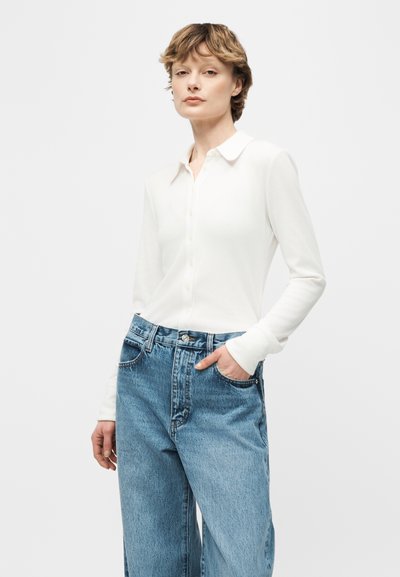 Γυναίκα με κοντά ανοιχτό καστανά μαλλιά, ντυμένη με λευκό πουκάμισο με γιακά και κλειστά κουμπιά και ψηλόμεσο μπλε τζιν, στέκεται με το ένα χέρι στην τσέπη.