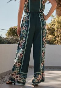 Bloemige jumpsuit van donkergroen stof, met een wijde pijp en een elastische tailleband. Accenten van lichtblauwe strepen en kleurrijke bloemenpatronen.