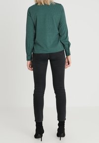 Grönrutigt långärmat skjorta som bärs med svarta jeans, med en avslappnad passform och knäppta manschetter, sedd bakifrån.