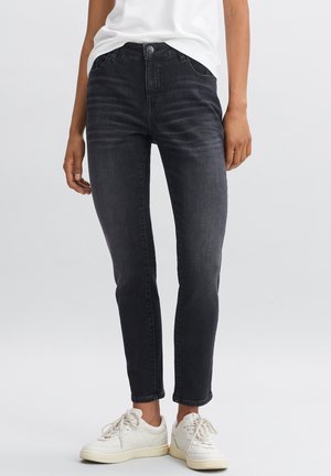 Person trägt schwarze Slim-Fit-Jeans und weiße Sneaker, steht vor einem einfarbigen hellgrauen Hintergrund.