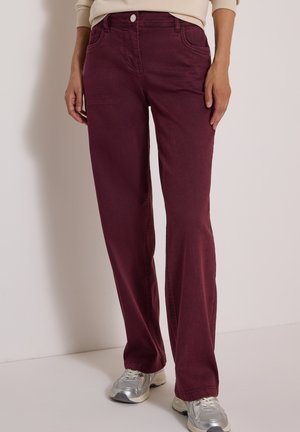 Personne portant un pantalon droit rouge foncé et des baskets argentées, debout devant un fond clair uni.