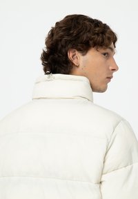 Chaqueta de plumas blanca con un alto cuello, textura acanalada y costuras en contraste. Presenta un cordón de ajuste en el cuello.