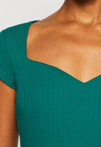 Prążkowana zielona sukienka z krótkimi rękawami, z kwadratowym dekoltem zwężającym się w delikatny kształt litery V na środku. Gładka faktura.