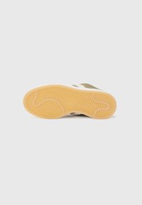Semelle de chaussure de sport en marron clair avec un motif de traction en zigzag. La tige est en toile verte avec des rayures blanches. La semelle en caoutchouc offre une bonne adhérence.