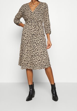 Kobieta ubrana w beżową i czarną sukienkę kopertową w panterkę z rękawami do łokci oraz czarne botki za kostkę.