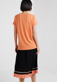 Kortärmad, orange T-shirt med rund hals, bärs tillsammans med en svart plisserad kjol med en orange och vit randig nederkant.