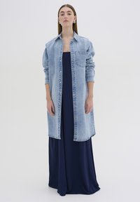 Giacca in denim azzurro chiaro con colletto e tasca frontale, sovrapposta a un abito lungo blu navy, tessuto liscio, design semplice.