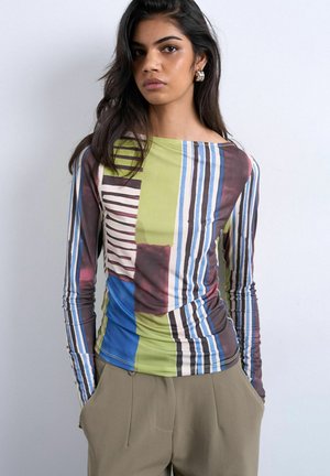 Ung kvinde med langt mørkt hår iført en flerfarvet, stribet langærmet top og beige bukser med hænderne i lommerne.