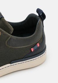 Green and navy slip-on sneakers featuring textured fabric, ribbed accents, beige rubber sole, and a decorative red, white, and blue tag.