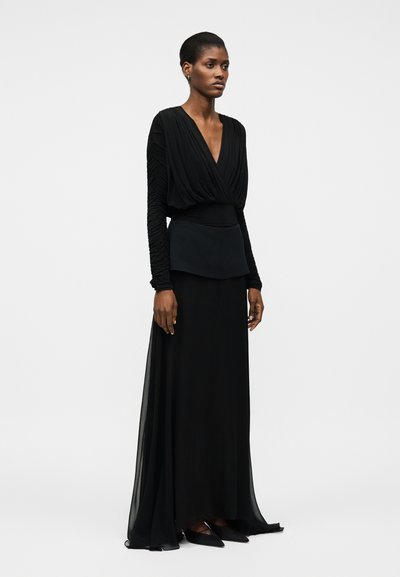 Kobieta w długiej czarnej sukni z marszczonymi długimi rękawami, głębokim dekoltem w kształcie litery V, dopasowaną talią i zwiewną spódnicą, stojąca na jednolitym tle.