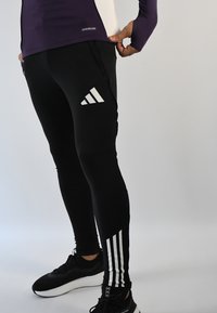 Leggings deportivos negros con un ajuste ceñido, que presentan logotipos blancos de Adidas y tres franjas blancas a lo largo de la parte inferior de las piernas. Textura suave, tela elástica.