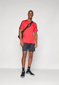 Červené sportovní tričko s černými pruhy, doplněné tmavými šortkami s texturovaným vzorem. Černé tenisky a malá černá taška přes rameno.