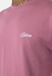 Nahaufnahme einer Person, die ein staubiges rosa T-Shirt mit einem weißen, geschwungenen "Situm"-Logo auf der Brust trägt.