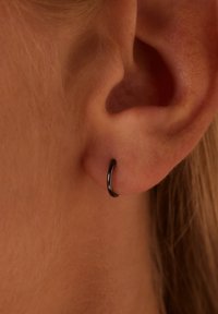 Zwarte metalen oorring getoond op een oorlel, met een glad, rond ontwerp en een dunne, gebogen silhouet met een gepolijste afwerking.