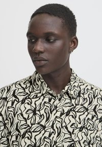 Short-sleeve button-up shirt in cream with a black leaf pattern. Features a collar, pocket on the left, and lightweight cotton material.