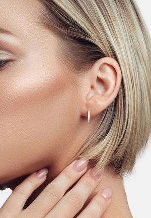 Sølv hoop ørepynt båret på en models øre; kort blond hår; glat hud med et naturligt makeup-look; minimalistisk design, reflekterende overflade.