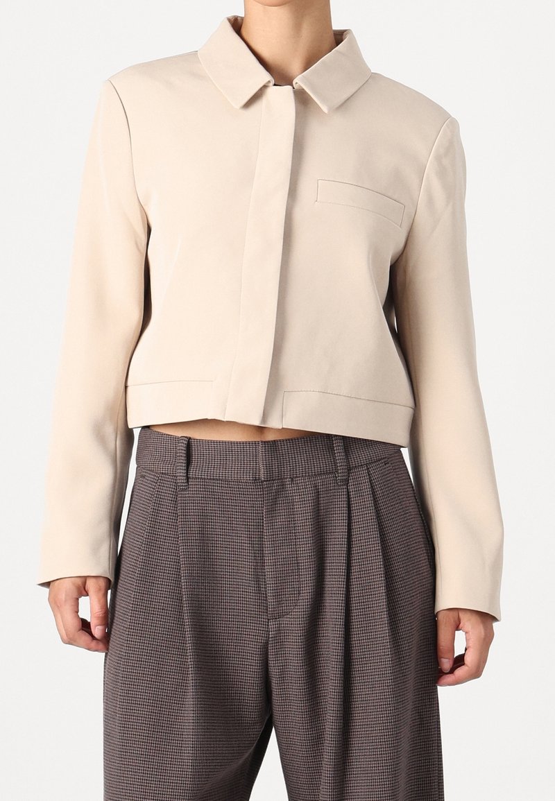 Veste courte beige avec boutons cachés, col cranté et poche poitrine, associée à un pantalon plissé à carreaux gris taille haute.