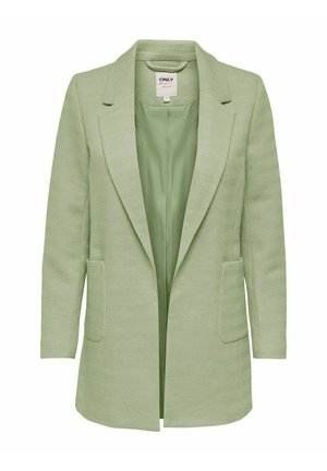 Nainen, jolla on vaaleanvihreä pitkä bleiseri, valkoinen neulepusero, mustat kapeat farkut ja mustat nilkkurit korolla, seisomassa yksinkertaisen taustan edessä.