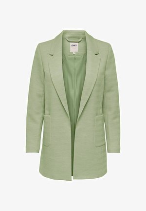 Vrouw die een lichtgroene lange blazer draagt, een witte gebreide trui, zwarte skinny jeans en zwarte enkellaarsjes met hak, staand tegen een effen achtergrond.