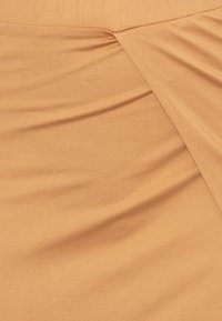 Tejido marrón claro con costura visible y ligero pliegue recogido, mostrando textura y puntadas en el material suave.