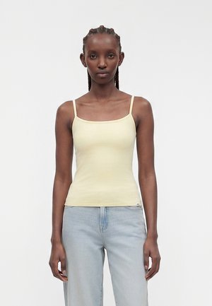 Kvinde med fletninger iført en lysegul spaghetti-strop top og lyseblå jeans, stående mod en ensartet lys baggrund.