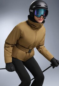 Brown ski jacket with a high collar, belt detail, and zippered sleeves, paired with black ski pants and gloves, worn with a black helmet and goggles.