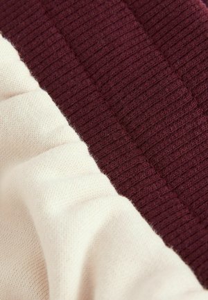 Frammento di maglione lavorato a maglia con una sezione a coste di colore bordeaux e una sezione liscia di colore crema, evidenziando materiali e colori a contrasto.