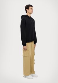 Felpa nera con cappuccio e vestibilità rilassata, abbinata a pantaloni cargo beige con molte tasche, indossata con sneakers bianche.