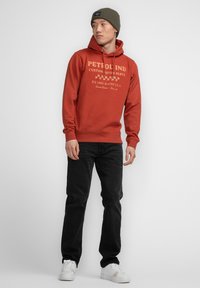 Felpa rossa con grafica testuale, coulisse e tasca frontale, abbinata a jeans neri e sneakers bianche; uomo che indossa un berretto verde.