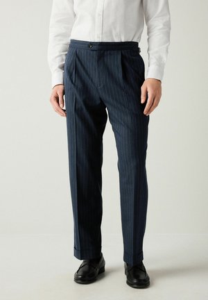 Pantalon rayé bleu foncé avec une coupe ajustée, plis, fermeture à bouton et poches latérales, fabriqué à partir d'un tissu lisse. Accompagné de mocassins noirs.