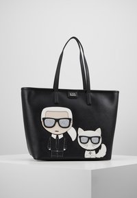 Zwarte tote bag met een cartoonontwerp van een figuur en een kat, beide met een zonnebril. Gemaakt van synthetisch materiaal met textuurelementen.