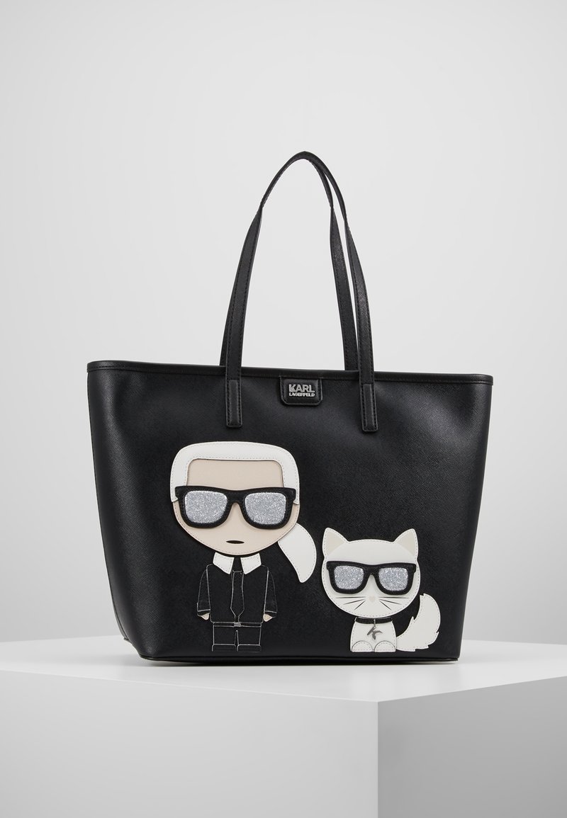 Zwarte tote bag met een cartoonontwerp van een figuur en een kat, beide met een zonnebril. Gemaakt van synthetisch materiaal met textuurelementen.