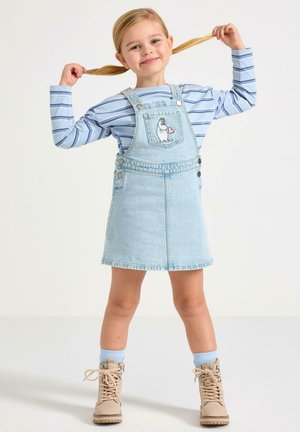 Denim tuinbroekjurk in lichtblauw, met een voorzak met een karakterprint, gecombineerd met een blauw-wit gestreept shirt met lange mouwen en beige laarzen.