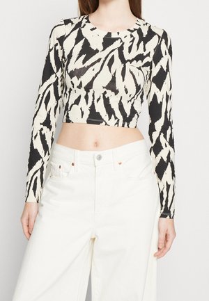 Nainen, jolla on mustavalkoinen abstrakti kuvioitu pitkähihainen lyhyt paita ja korkea vyötäröiset valkoiset leveälahkeiset housut.